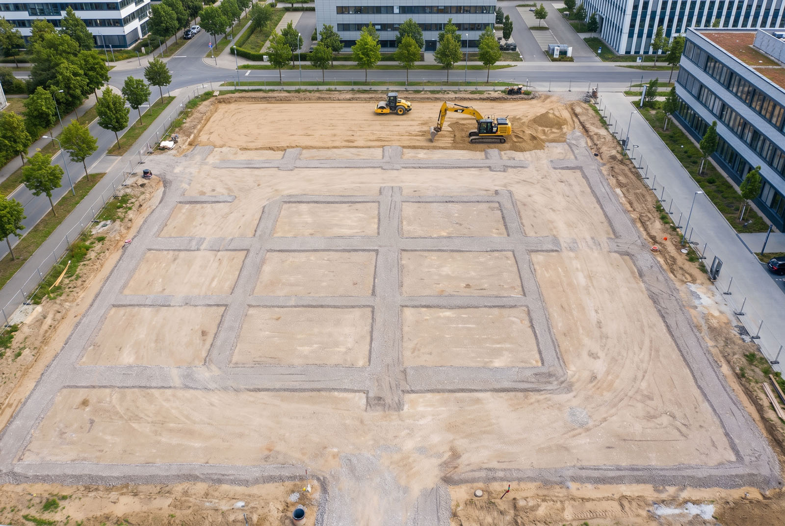 Widok z drona na plac budowy z precyzyjnie wykonanymi wykopami pod ławy fundamentowe i ciężkim sprzętem w tle.